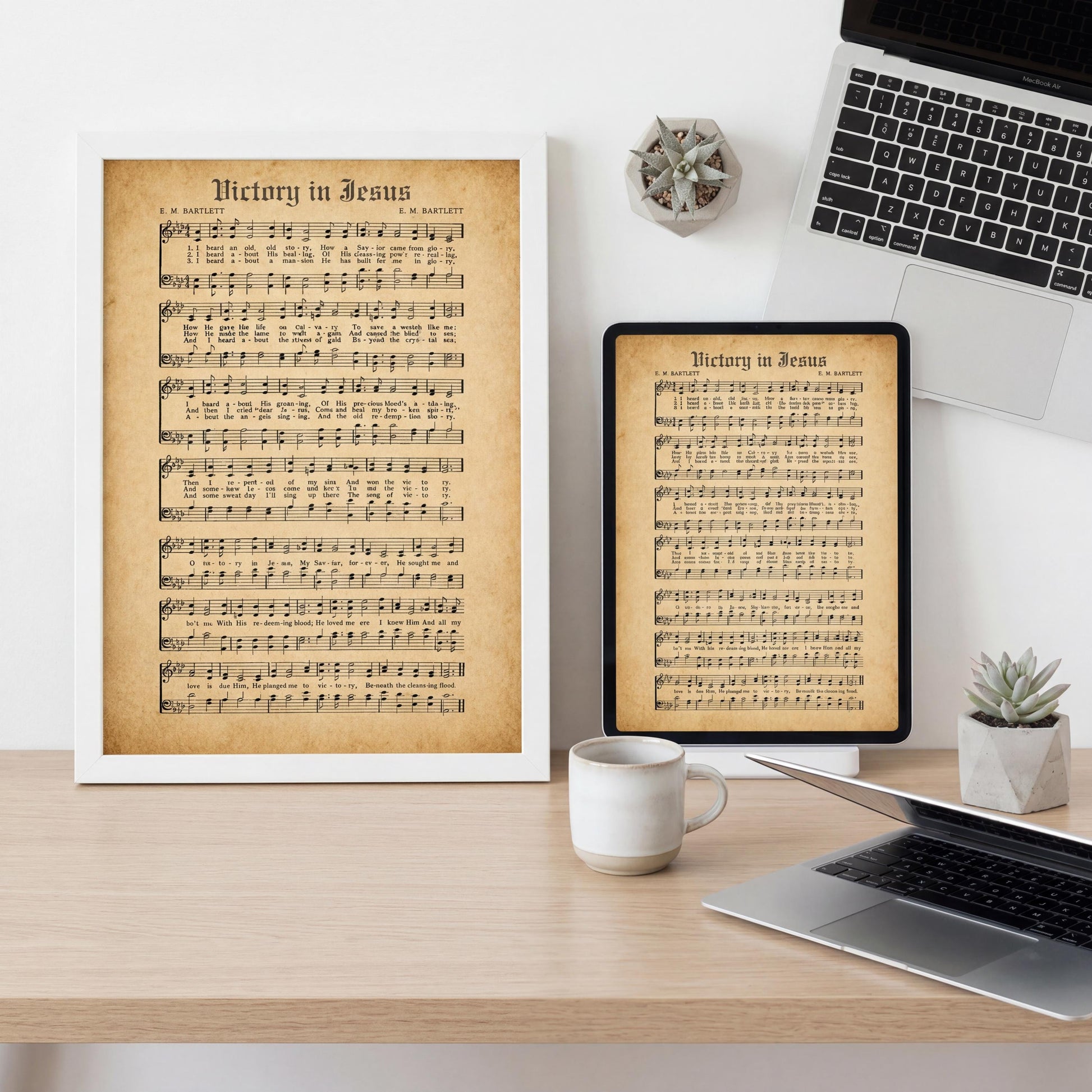 a wooden desk with a framed sheet of music titled "Victory in Jesus" and a laptop, a coffee cup, and a small potted plant.