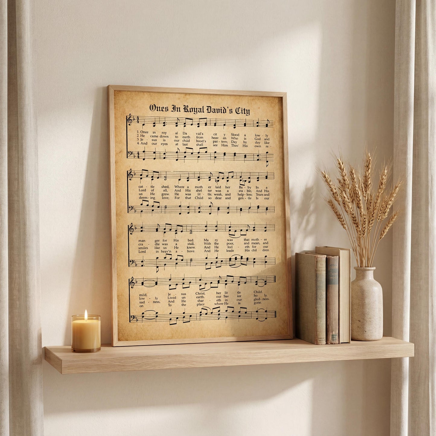 a framed sheet music poster of a hymn titled "Once in Royal David's City" displayed on a wooden shelf, accompanied by a lit candle, a vase of wheat stalks, and a stack of books.