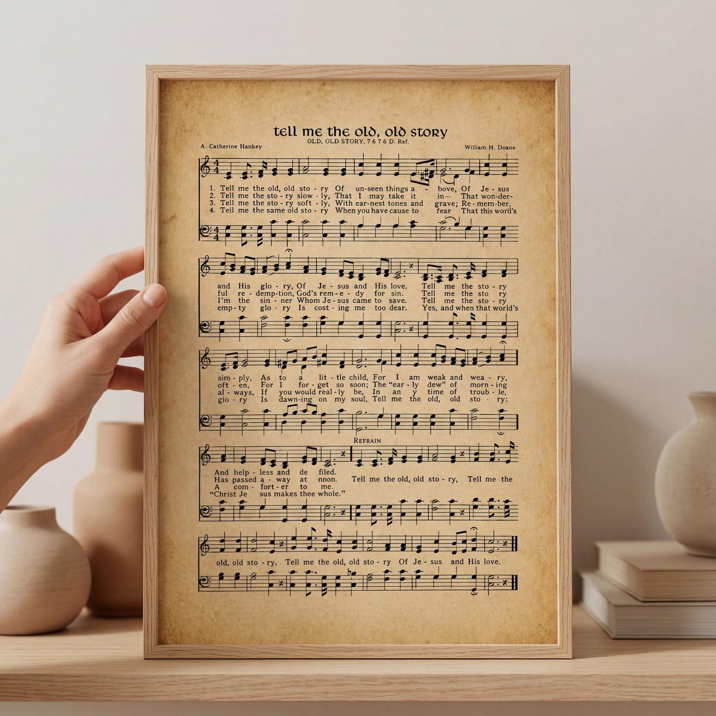 A framed sheet of music titled "tell me the old, old story" is being held up against a white wall, with a hand visible on the left side of the frame.