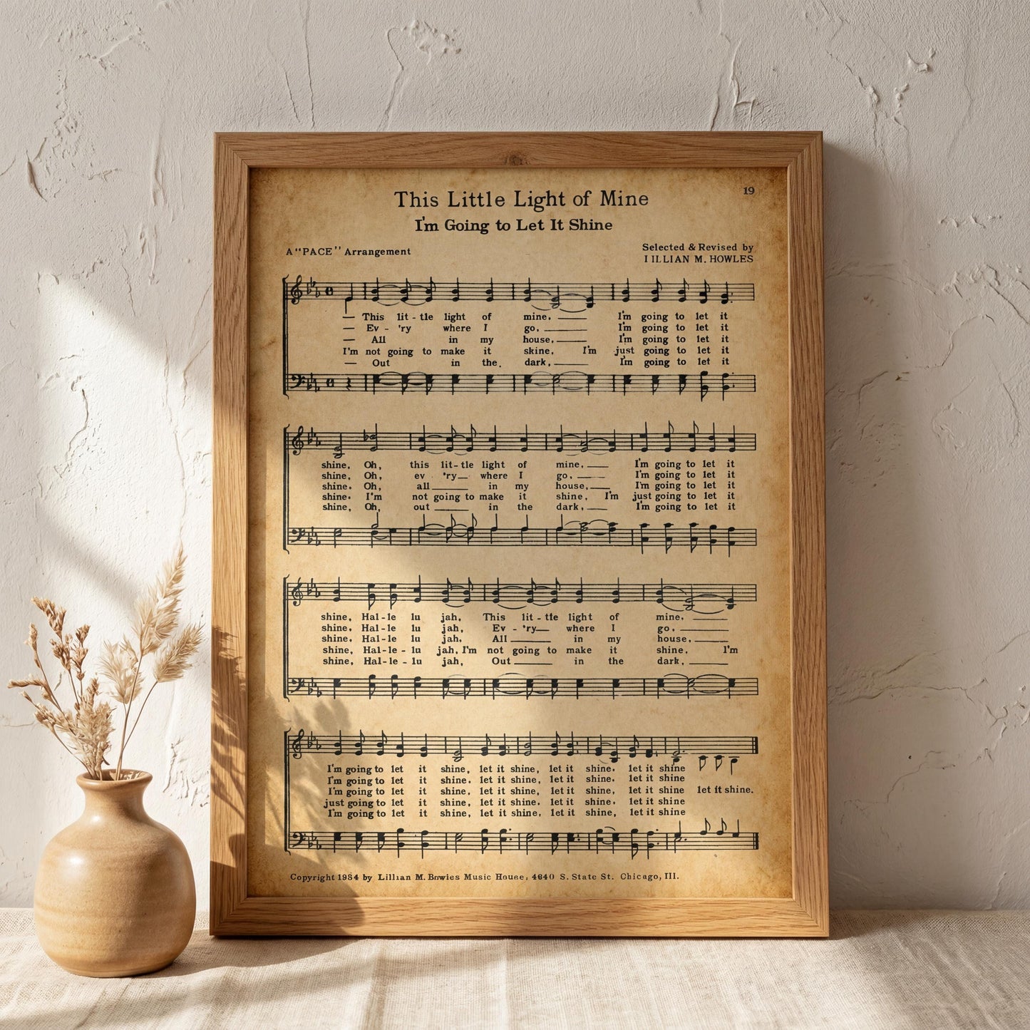a framed sheet music print of a song titled "This Little Light of Mine" arranged by Iliian &owesby. The sheet music is displayed on a wooden stand, with a vase of dried flowers placed next to it.