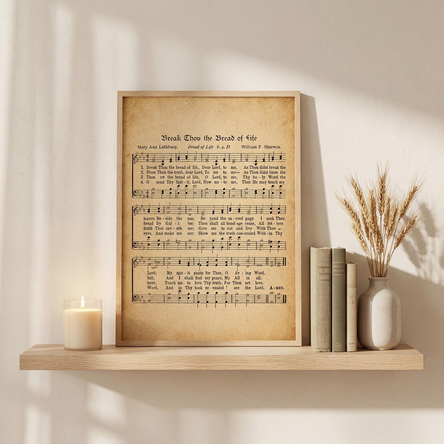a framed sheet music print of a hymn titled "Break The Bread of Life" displayed on a wooden shelf, accompanied by a lit candle, some books, and a vase of wheat stalks.