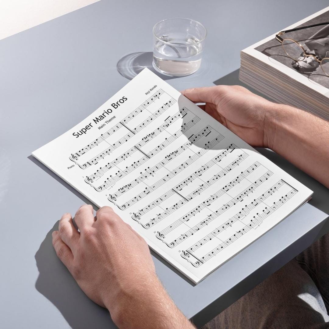 Printed Super Mario Bros. sheet music is shown held in hand at a desk setup with personal items like glasses and water, suggesting the piece’s use in casual practice or music education settings.