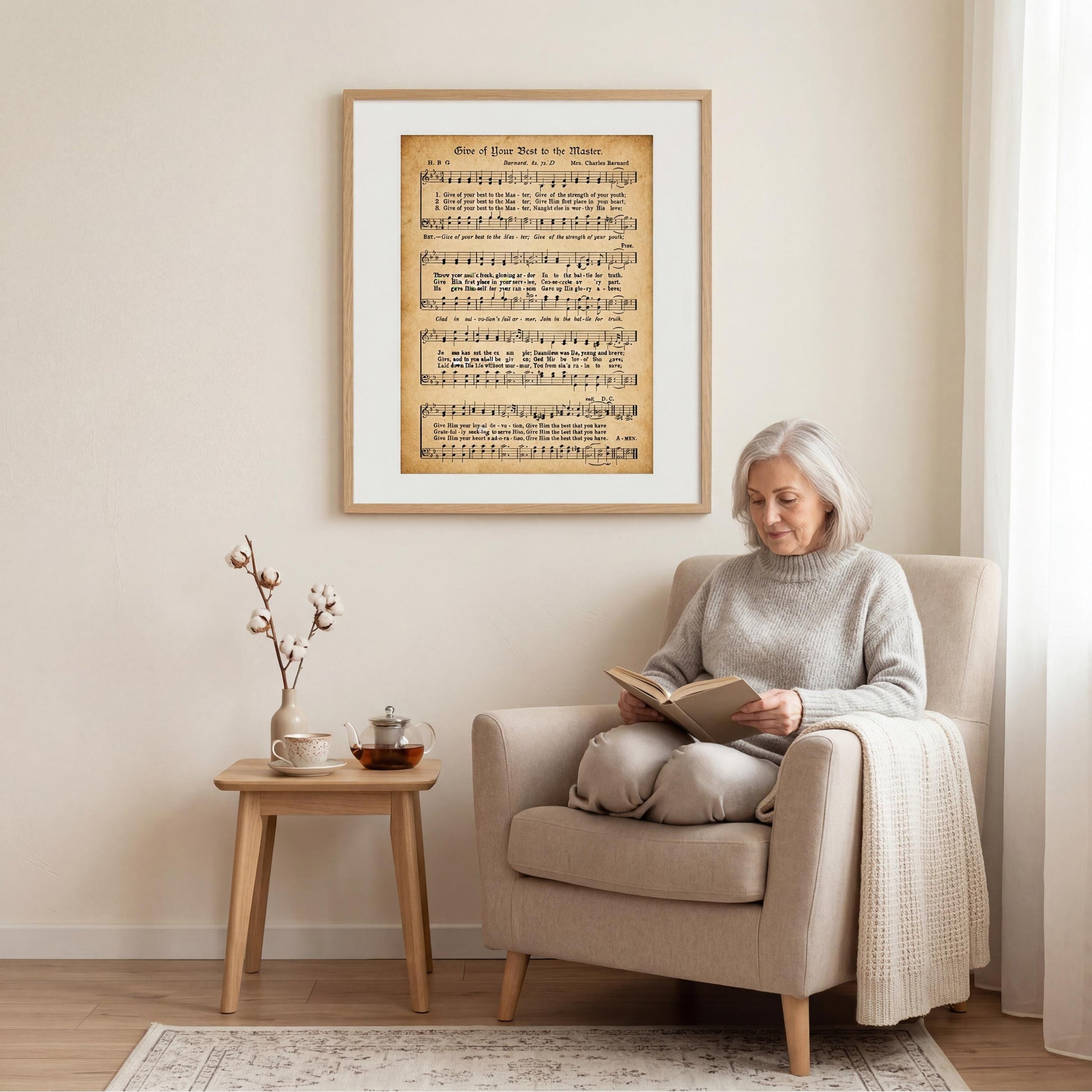 A woman is sitting in a beige armchair, reading a book, with a framed sheet music poster of a musical score hanging on the wall behind her.