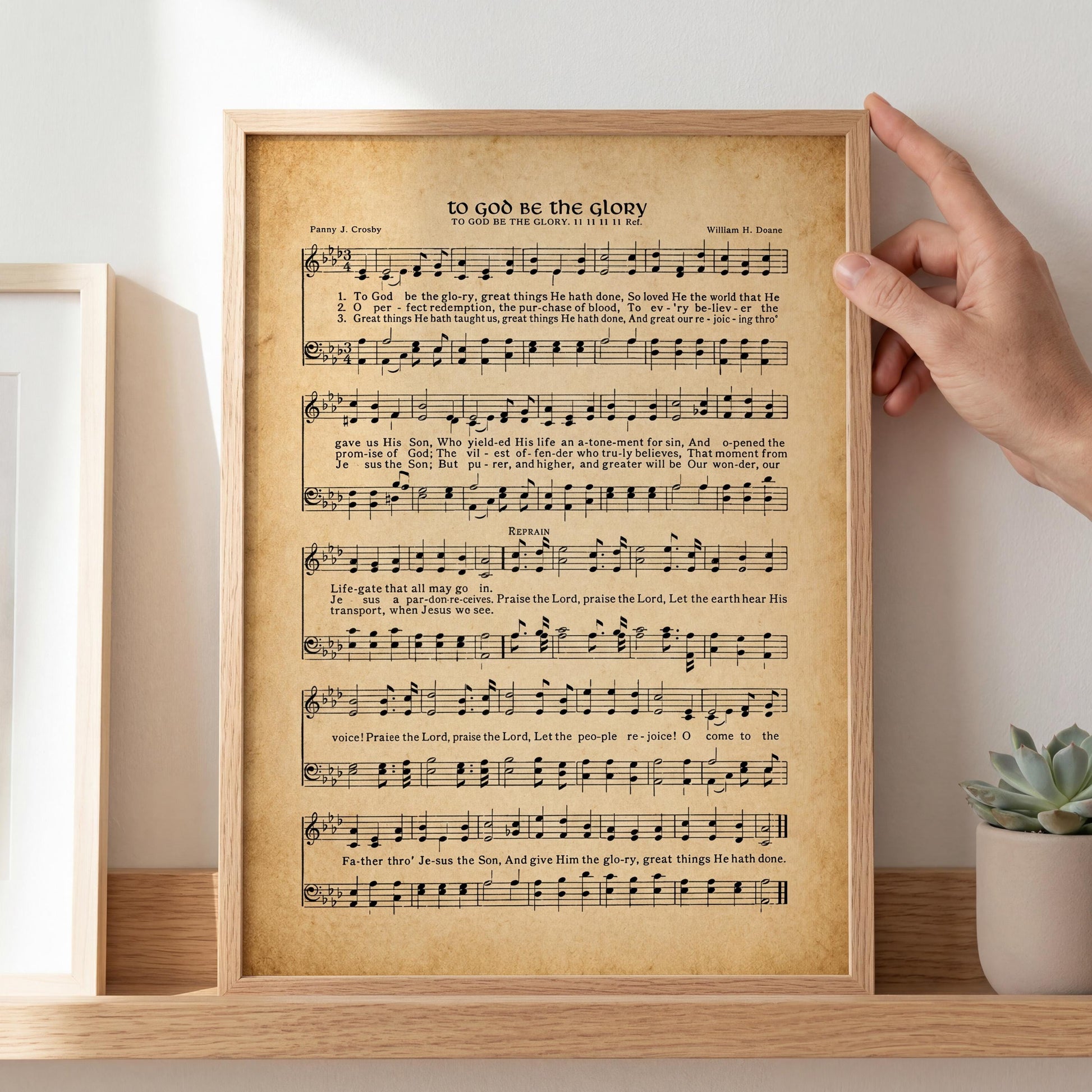 A framed sheet music print titled "To God Be the Glory" is displayed on a shelf, with a hand resting on it.