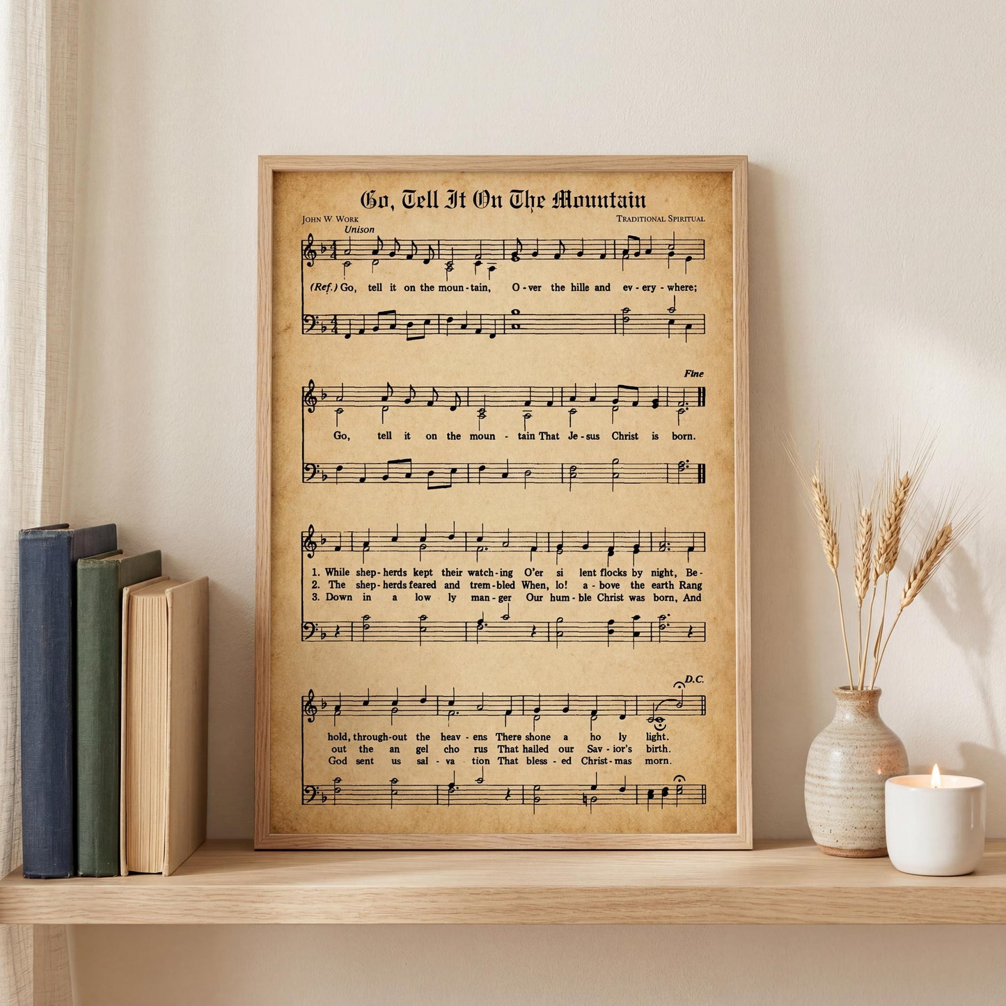A framed sheet music print titled "Go, Tell It On The Mountain" is displayed on a shelf next to a stack of books and a vase with wheat stalks.