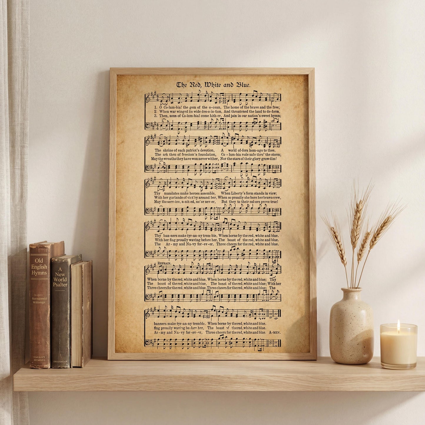 a framed sheet music print of "The Red White and Blue Hymn" displayed on a shelf, accompanied by a vase with wheat stalks and a lit candle.