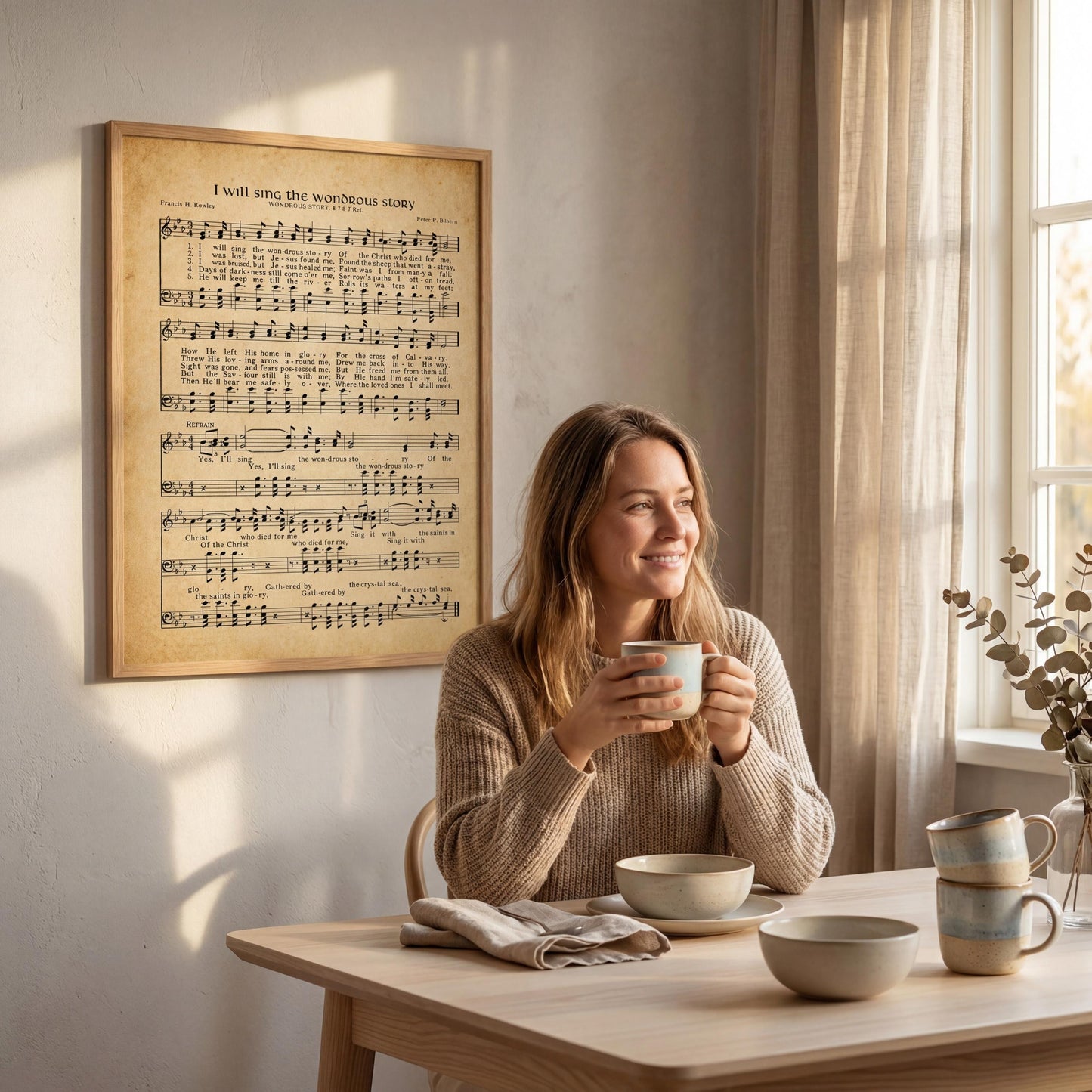 A woman is sitting at a table, holding a cup and smiling, with a framed sheet music poster on the wall behind her.