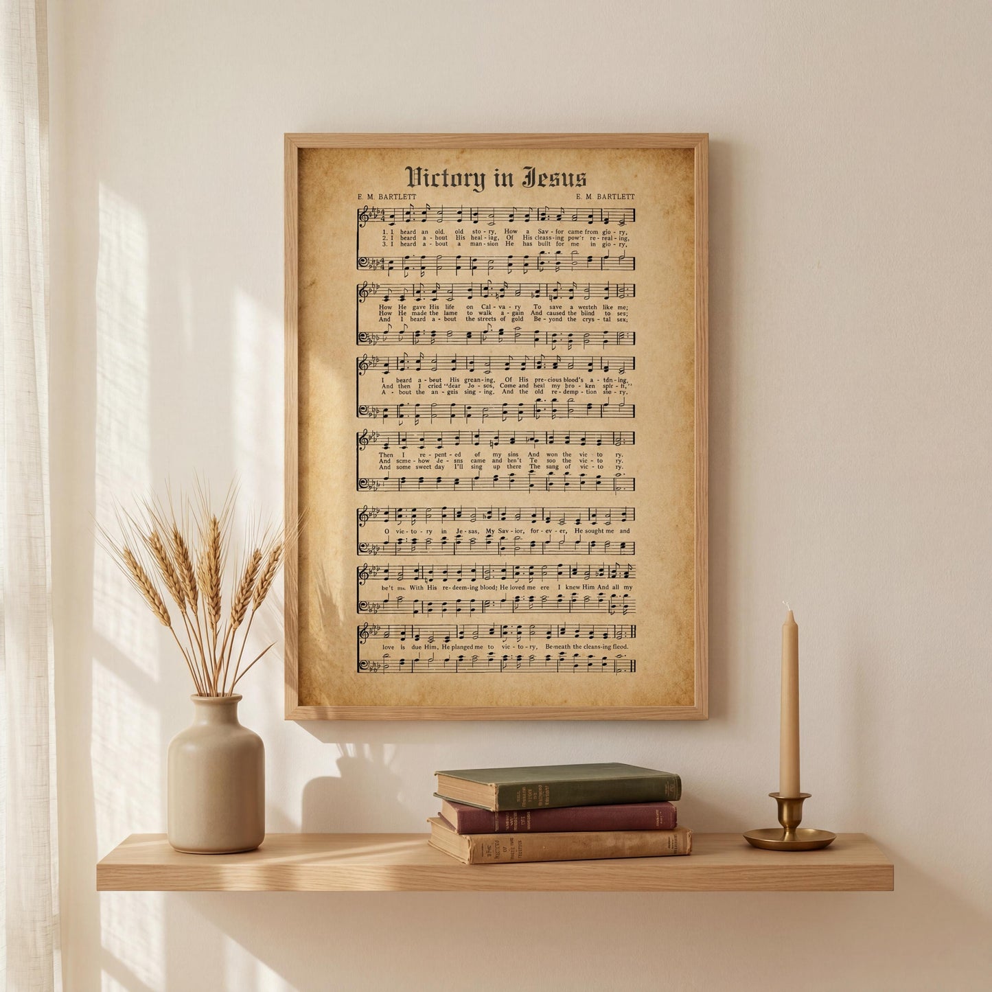 a framed sheet music poster of "Victory in Jesus" displayed on a wall above a wooden shelf with a vase of wheat stalks and a stack of books.