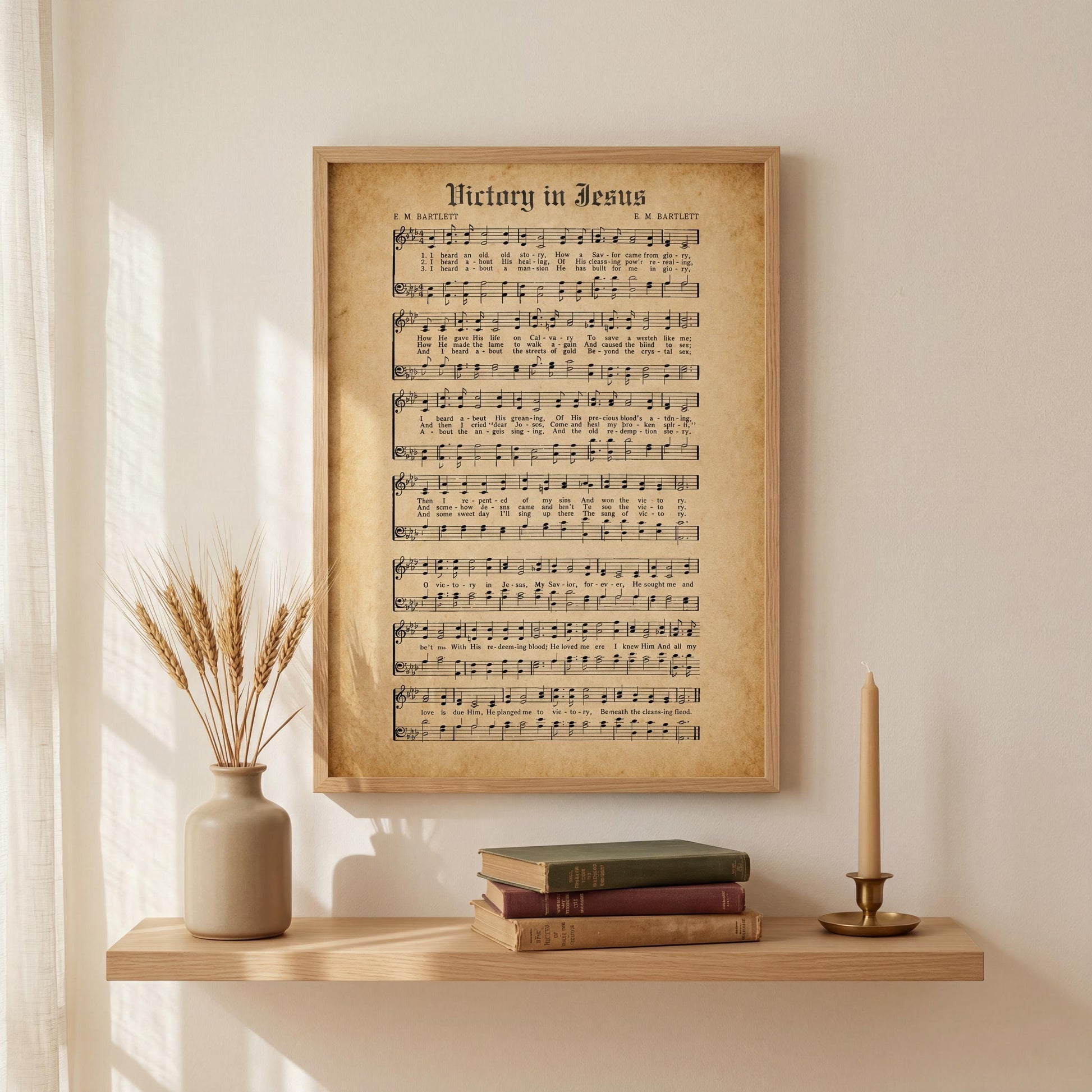 a framed sheet music poster of "Victory in Jesus" displayed on a wall above a wooden shelf with a vase of wheat stalks and a stack of books.