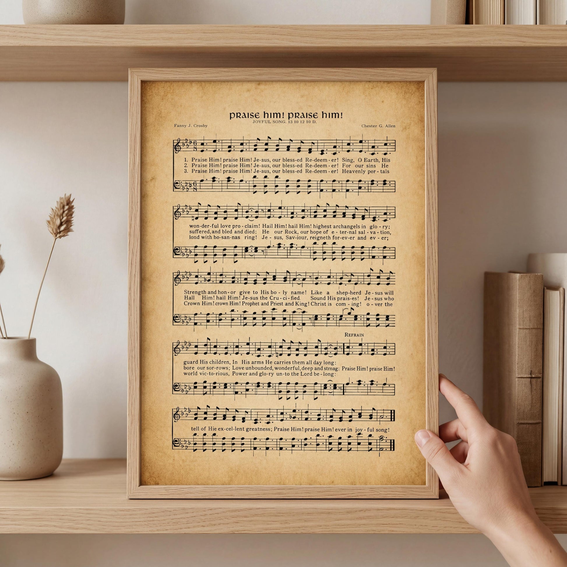 A framed sheet of music titled "Pray him, praise him!" is being held up by a hand, with a shelf of books and a vase of flowers in the background.