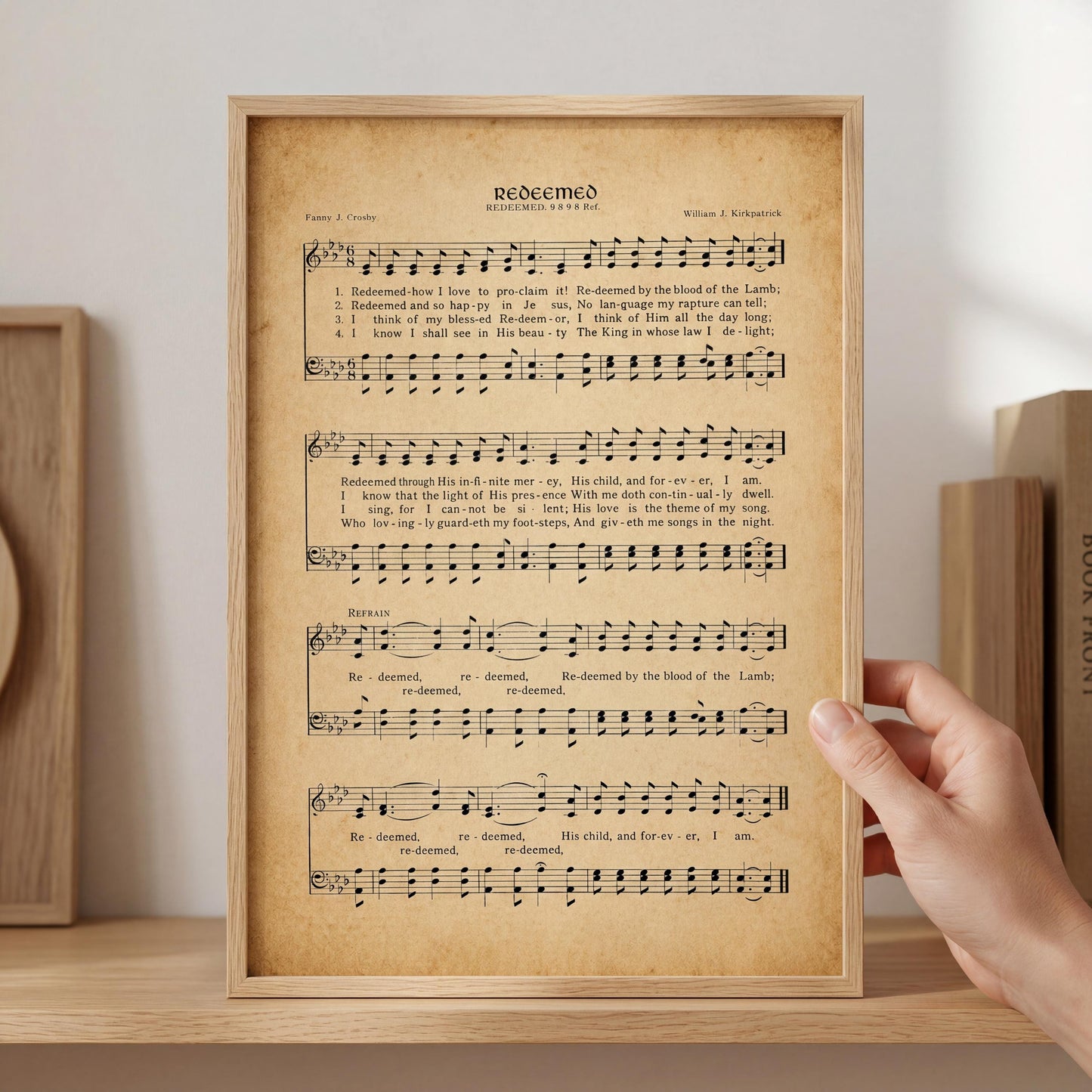 A framed sheet of music titled "Redeemed" is being held by a person's hand, with a wooden shelf and other books visible in the background.